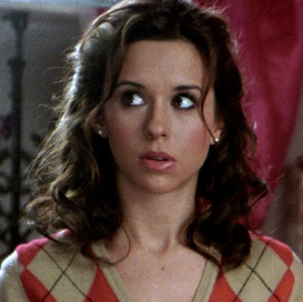 lacey chabert, a white feminine person with medium length dark brown hair, brown eyes and a shirt with an argyle diamond pattern is staring off camera neutrally. the background is unclear and blurry.