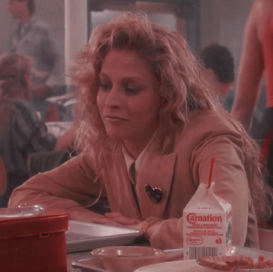 lissane falk, a white feminine person with messy long blonde hair, brown eyes and a yellow suit jacket is sitting at a lunch table and staring neutrally off camera. the background is a school caffateria.